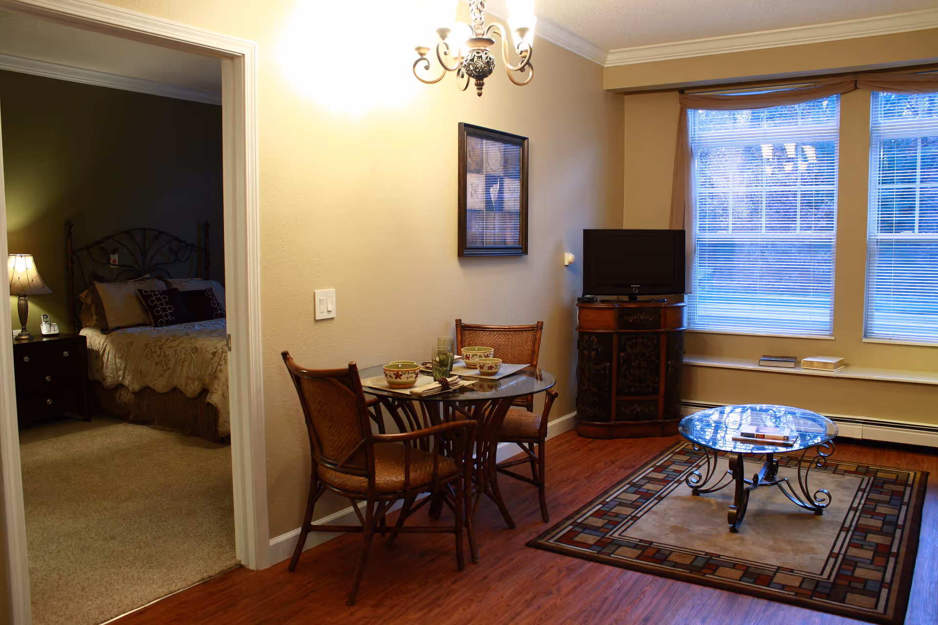 A cozy living area with a small round glass dining table set with two chairs and tableware. Adjacent to the dining area is a bedroom with a bed, nightstand, and lamp. The living area has a glass coffee table on a patterned rug, a small cabinet with a TV on top, and large windows with blinds letting in natural light.