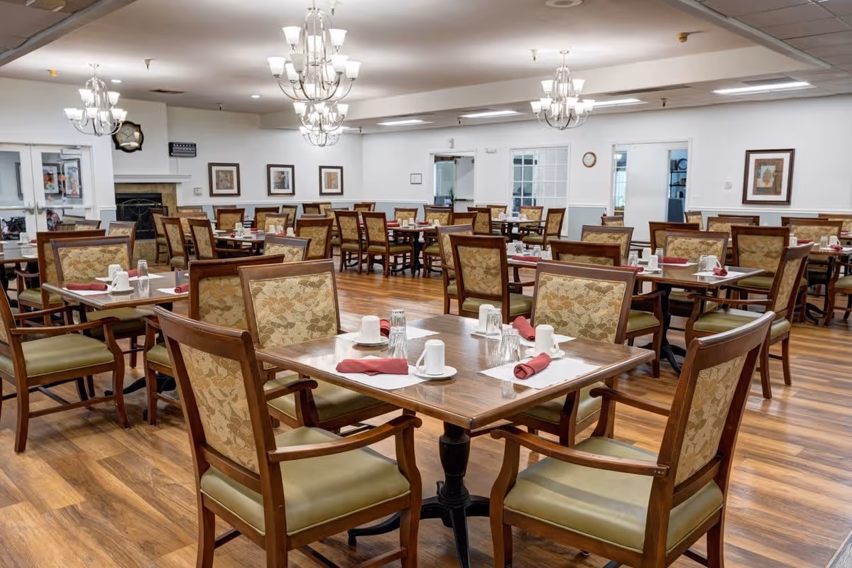 A spacious dining room with multiple wooden tables and cushioned chairs arranged neatly. Each table is set with white cups, glasses, plates, and red cloth napkins. The room has wooden flooring, white walls adorned with framed artwork, and several elegant chandeliers hanging from the ceiling providing warm lighting.
