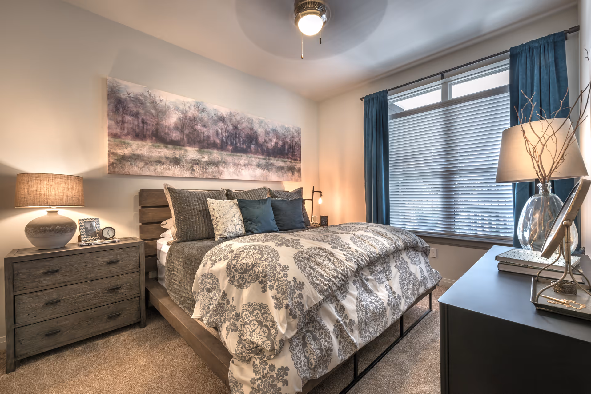 A cozy bedroom with a large bed featuring patterned bedding and multiple pillows. There is a wooden nightstand with a lamp, a photo frame, and a clock on the left side of the bed. A large window with blinds and blue curtains is on the right side, next to a black dresser with a lamp, books, and decorative items. A ceiling fan with a light is mounted on the ceiling, and a landscape painting hangs above the bed.