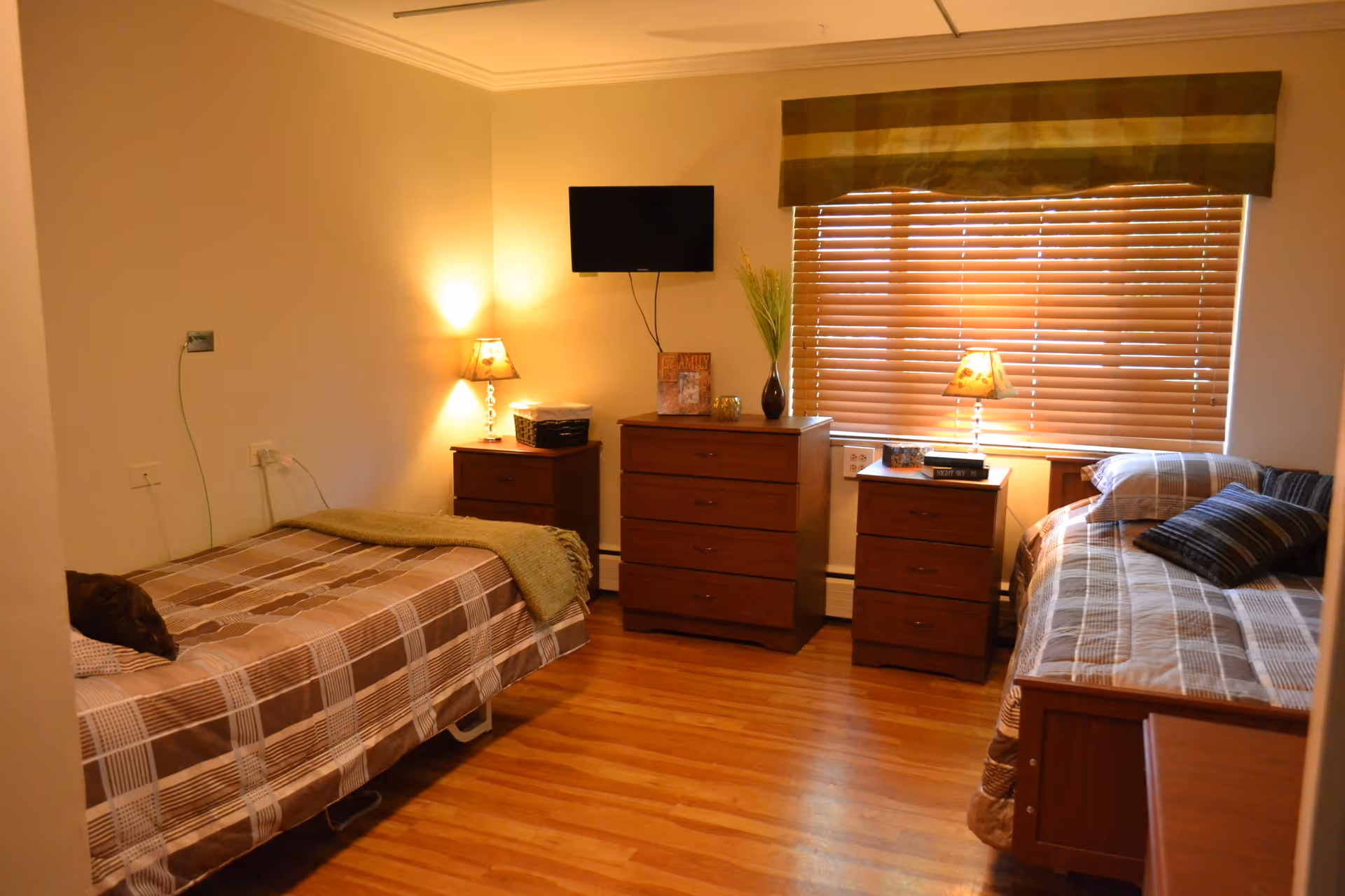 A cozy bedroom with two single beds on opposite sides of the room, each with brown and beige plaid bedding. Between the beds are three wooden dressers, two with table lamps and decorative items on top. A flat-screen TV is mounted on the wall above the middle dresser. The window has wooden blinds and a valance, and the floor is hardwood.
