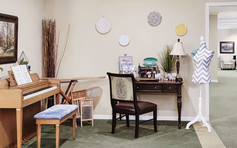 A cozy interior room featuring a wooden piano with a blue cushioned bench, a vintage washboard, a wooden desk with a chair, decorative embroidery hoops on the wall, a table lamp, a plant, and a dress form mannequin with a chevron pattern cover. The room has green carpet and beige walls, with an open doorway leading to another room with armchairs.