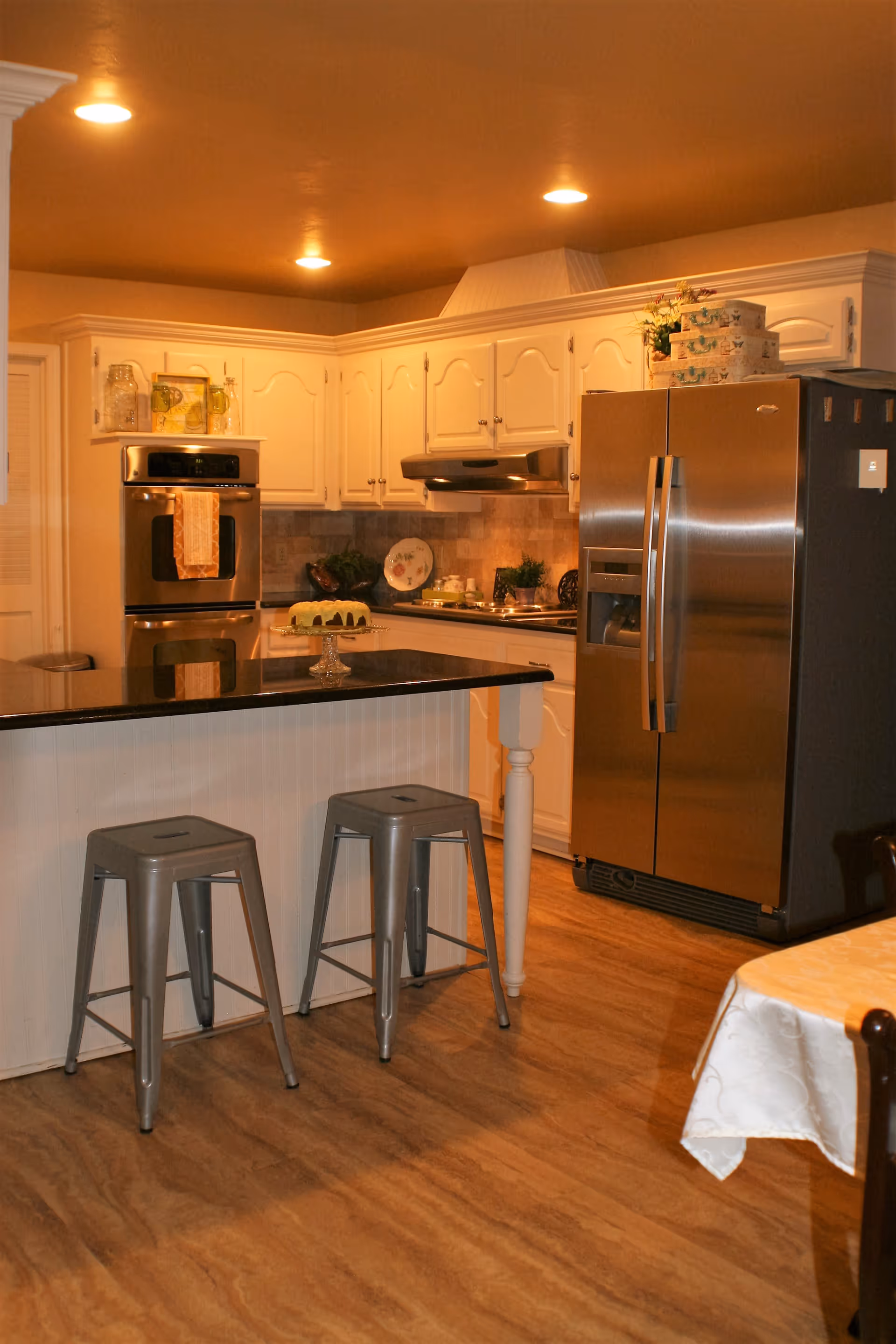 A warm, well-lit kitchen with white cabinetry and stainless steel appliances including a double oven and a large refrigerator. There is a black countertop island with two metal stools in front of it. A cake is displayed on a glass cake stand on the island. The floor has a wood-like finish, and a dining table with a white tablecloth is partially visible on the right side.