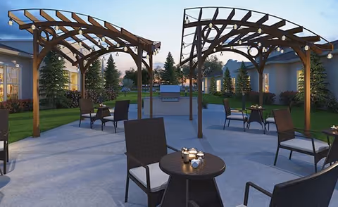 Outdoor patio area at a senior living facility with multiple wooden pergolas, several round tables with chairs, string lights hanging from the pergolas, and a grill in the background surrounded by greenery and trees during dusk.
