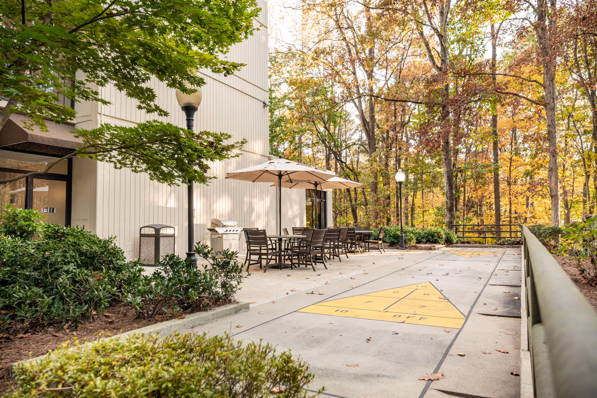 Outdoor patio area at Smyrna Towers Senior Apartments featuring a shuffleboard court, several tables with chairs under large umbrellas, a barbecue grill, surrounded by trees with autumn foliage.