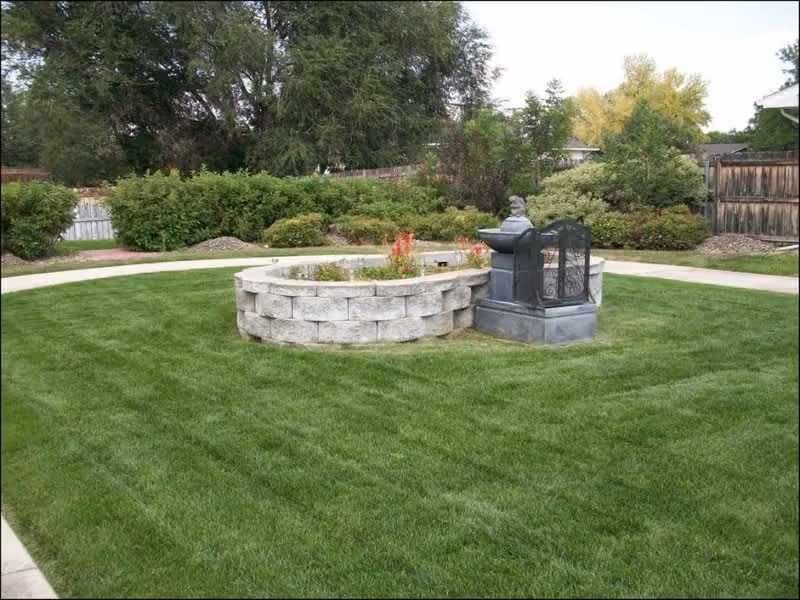 A grassy courtyard with a circular raised stone planter and a decorative fountain, surrounded by shrubs and trees.