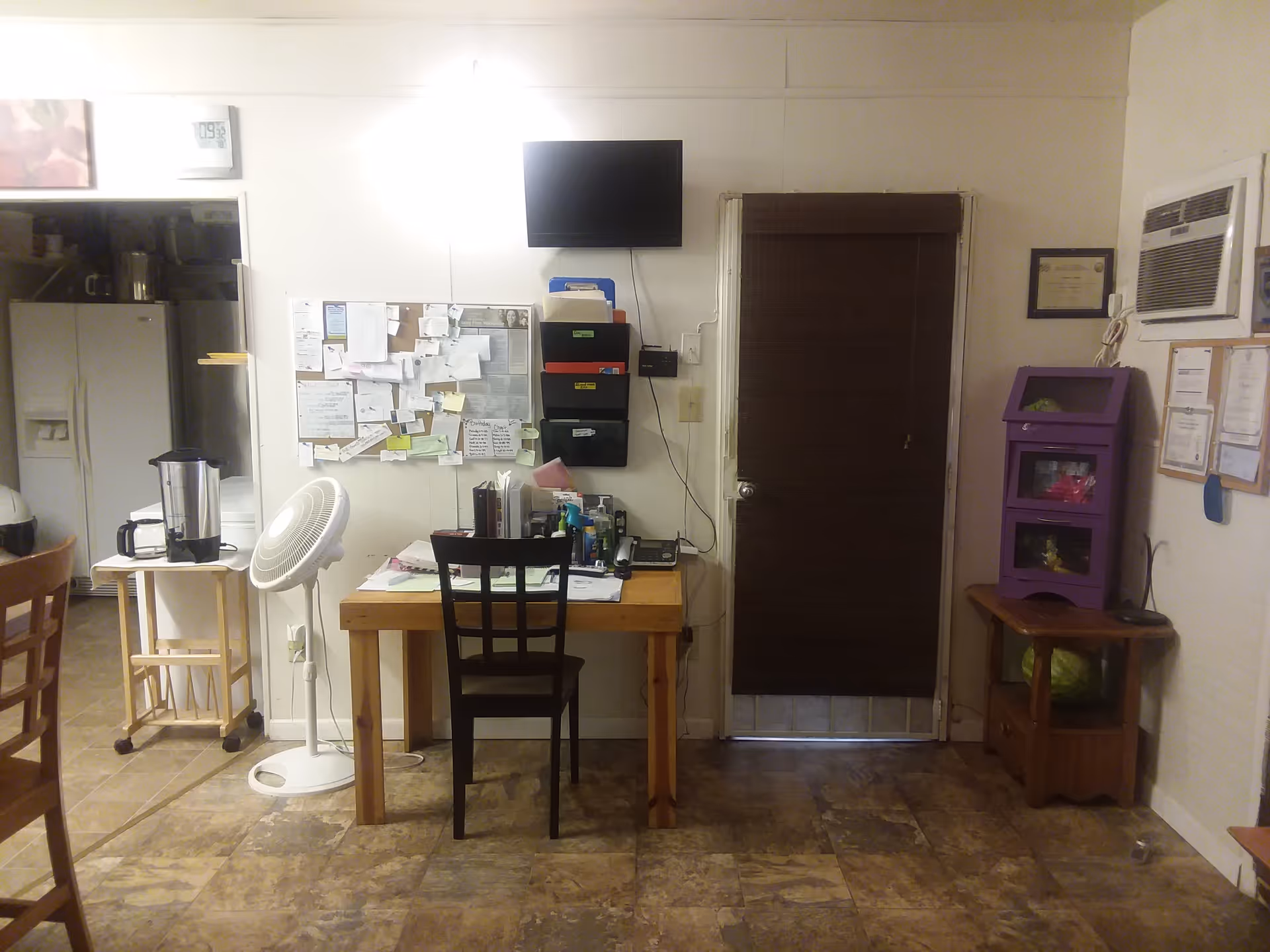 Small communal room with a desk and chair in front of a bulletin board and wall-mounted TV, a door with a closed blind, and a view into a kitchen area.
