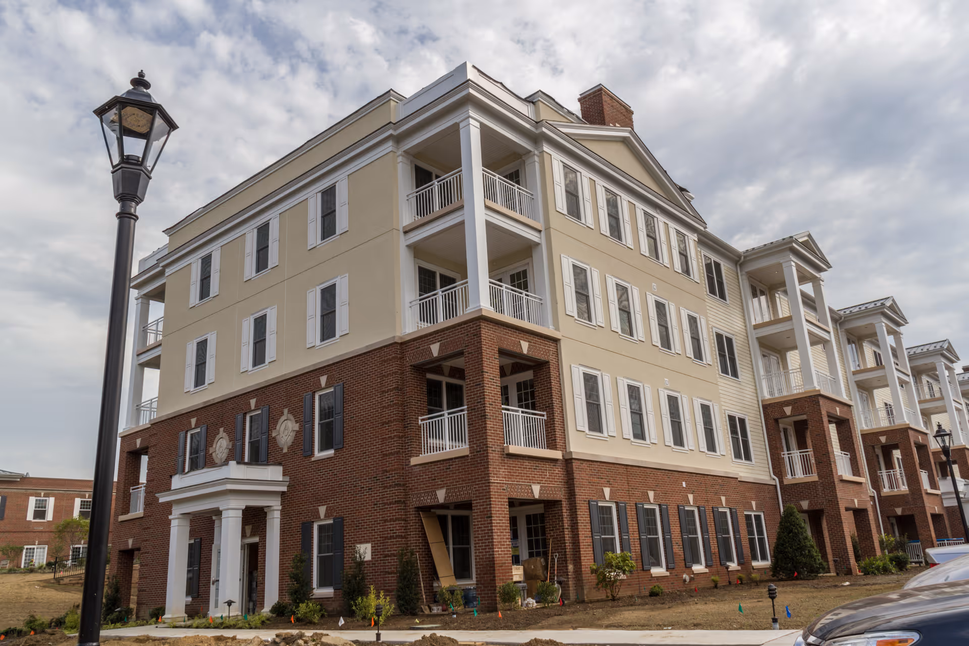 Exterior view of a multi-story residential building with brick and beige siding, white balconies, and multiple windows under a cloudy sky. A black street lamp is visible in the foreground.