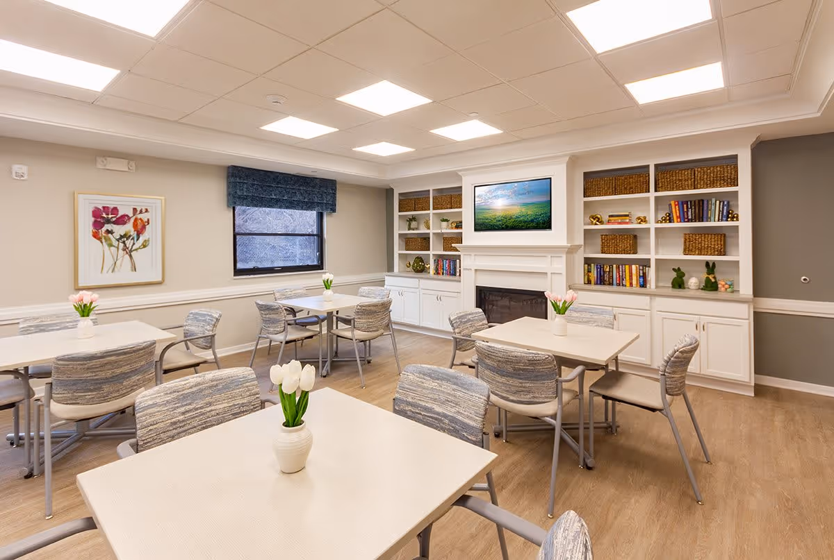 A bright and clean dining room with several tables and chairs arranged neatly. Each table has a small vase with white tulips. The room features a window with a blue valance, a framed floral artwork on the wall, built-in white shelves filled with books and decorative items, and a fireplace with a TV mounted above it.