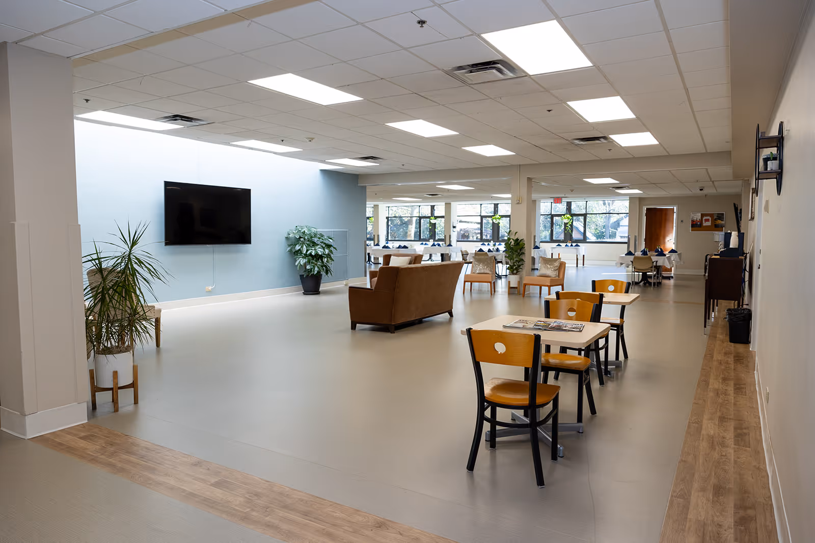 A spacious common area in a senior living facility with a mounted flat-screen TV on a light blue wall, several potted plants, a brown sofa, multiple wooden chairs and tables, and large windows letting in natural light.