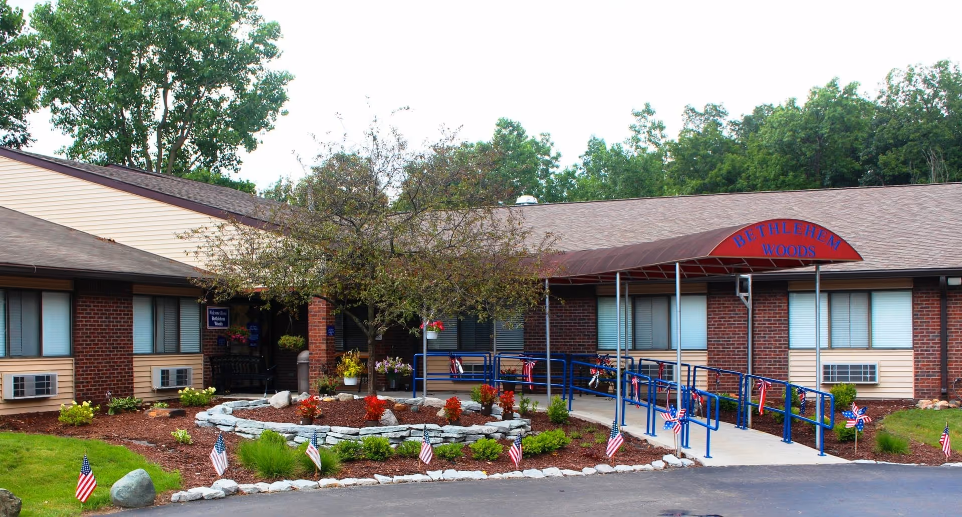 Front exterior of Bethlehem Woods Nursing and Rehabilitation with an entrance canopy, wheelchair ramp, landscaped garden and small American flags.