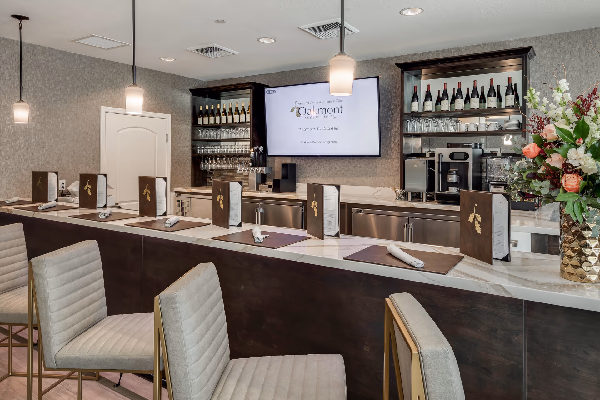 Interior view of a modern bar area in a senior living facility with a marble countertop, six bar stools with light gray upholstery, menus and napkins placed on the counter, shelves with wine bottles, glasses, and coffee machines in the background, and a large screen displaying the Oakmont Senior Living logo and slogan.