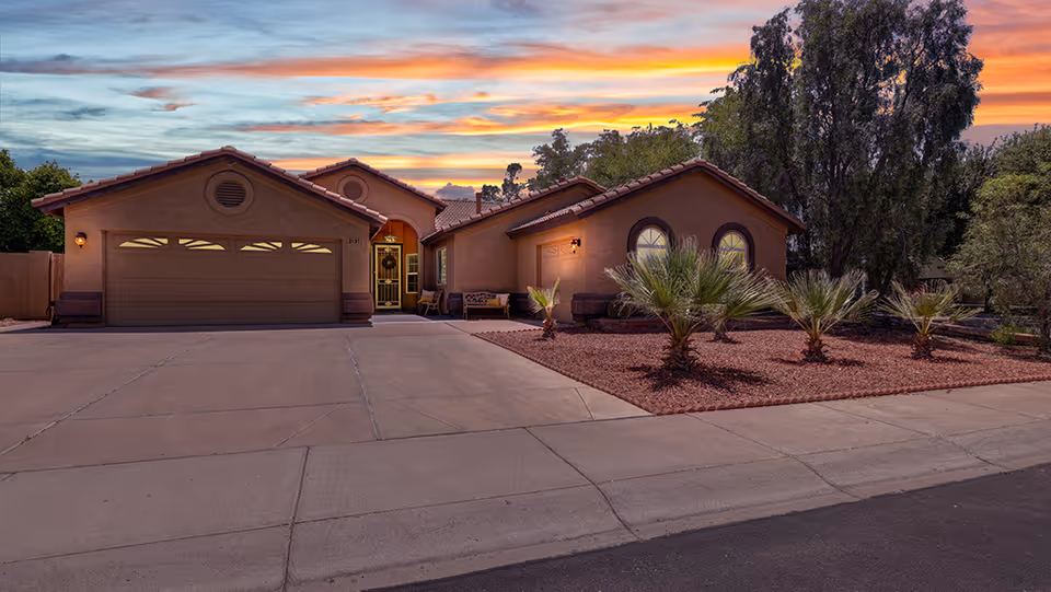 Single-story stucco home with a three-car garage, desert landscaping, and a colorful sunset sky.
