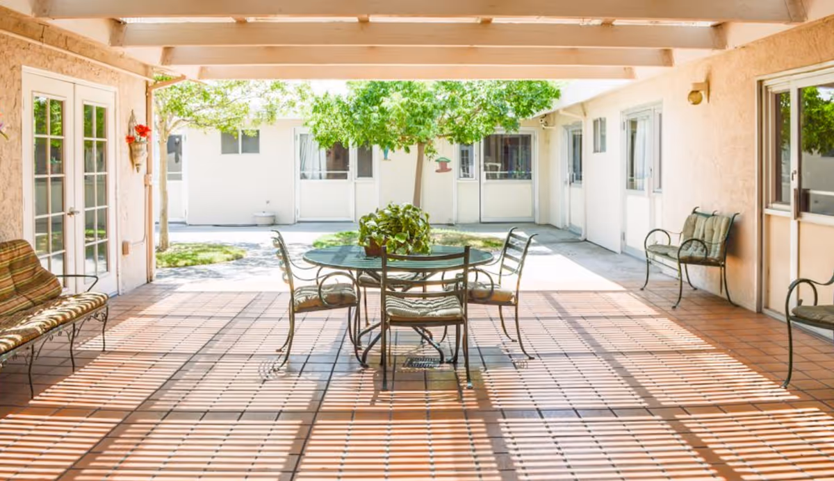 Covered outdoor patio area with a round glass table and four metal chairs with cushions. There are two cushioned benches on either side against the walls. The patio floor is tiled with terracotta tiles, and sunlight creates striped shadows from the overhead beams. In the background, there is a courtyard with green trees and white building walls with doors and windows.