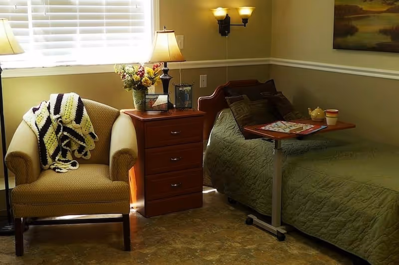 A cozy bedroom with a single bed covered in a green quilt, a wooden bedside table with a lamp, flowers, and picture frames, and a comfortable armchair with a crocheted blanket draped over it. A small overbed table holds a teapot, cup, and a magazine. The room has beige walls and a window with white blinds letting in natural light.