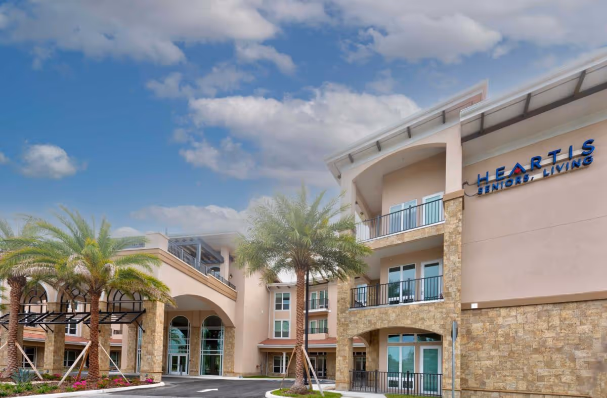 Exterior view of Heartis Venice Assisted Living building with palm trees, stone and stucco facade, balconies, and a covered entrance under a partly cloudy sky.