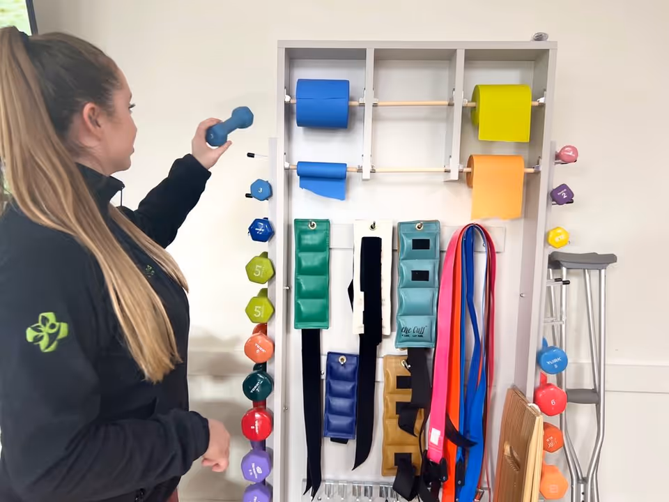 A woman with long hair tied in a ponytail is reaching for a blue dumbbell from a rack that holds various colorful dumbbells, resistance bands, and exercise weights. A crutch is leaning against the wall nearby.