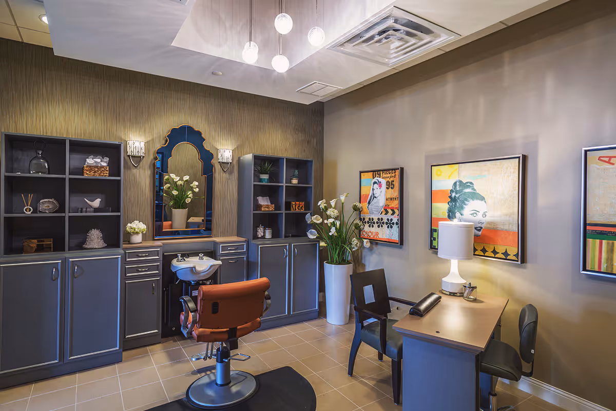 Interior view of a stylish salon room with a brown salon chair in front of a sink and mirror. The room features gray cabinets with shelves holding decorative items, a tall white vase with flowers, a desk with two chairs, a table lamp, and colorful framed artwork on the walls. The ceiling has modern pendant lights.