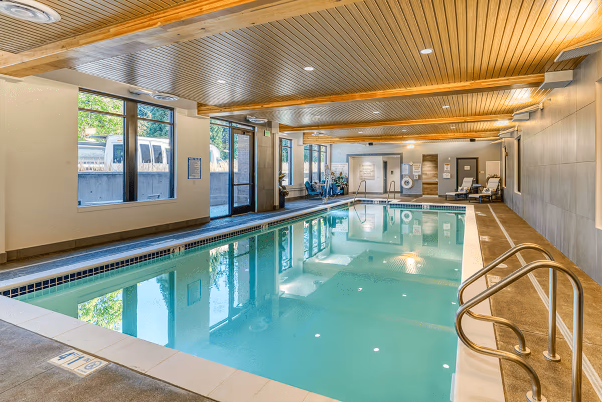 Indoor swimming pool area with clear water, surrounded by tiled flooring and walls. The ceiling has wooden paneling with recessed lighting. Large windows and glass doors on one side allow natural light to enter. Pool safety equipment and lounge chairs are visible at the far end of the pool.