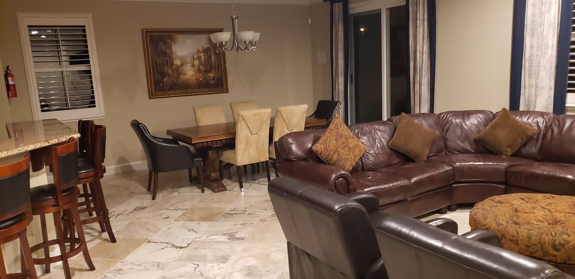 Interior view of a living and dining area featuring a brown leather sectional sofa with three brown cushions, a matching patterned ottoman, two black leather armchairs, a wooden dining table with six chairs, a chandelier above the table, a framed painting on the wall, and a kitchen counter with four wooden bar stools. The room has beige walls, marble flooring, windows with curtains, and a fire extinguisher mounted on the wall.