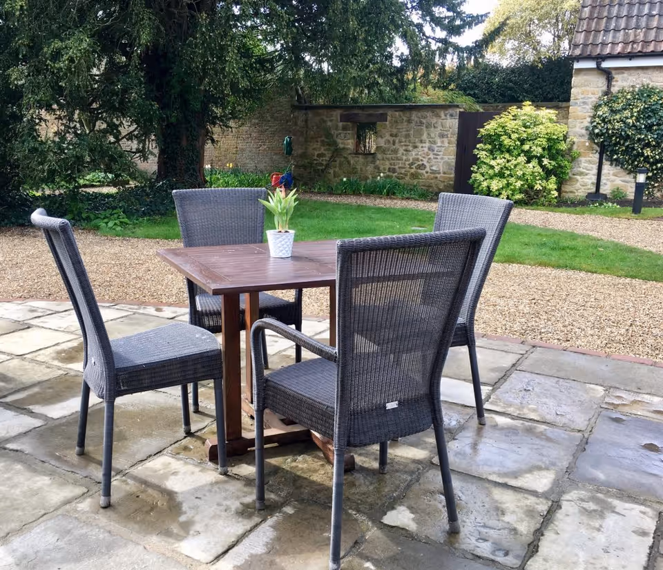Outdoor patio with four wicker chairs around a wooden table on a stone surface, with a lawn and stone wall in the background.