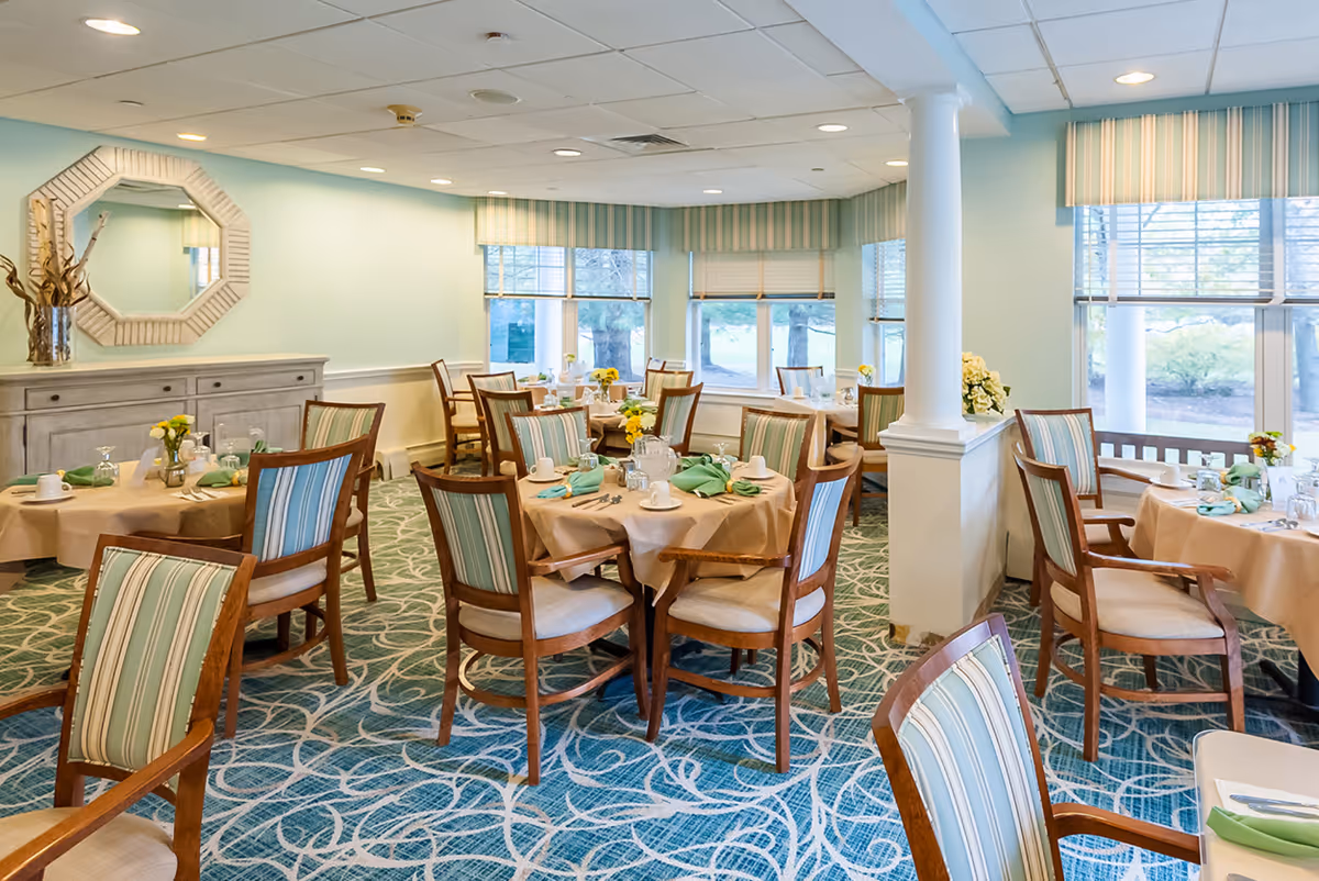 A bright dining room with round tables covered in beige tablecloths, each set with cups, glasses, silverware, and green napkins. The chairs have wooden frames with striped upholstery in shades of green and blue. Large windows with striped valances let in natural light, and a decorative mirror hangs on a light green wall above a sideboard with a vase of dried branches.