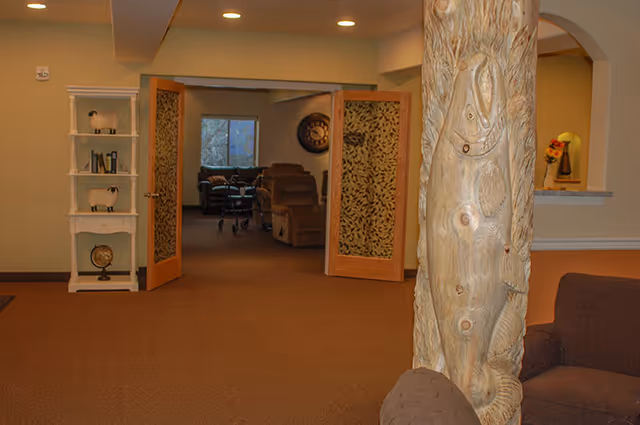Common seating area with a carved wooden column in the foreground and patterned double doors opening to a lounge with recliners.