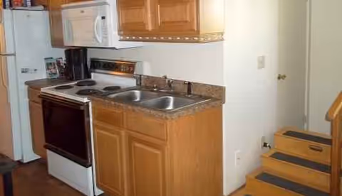 Compact kitchen with stove, sink, microwave, wooden cabinets, and nearby stairs.
