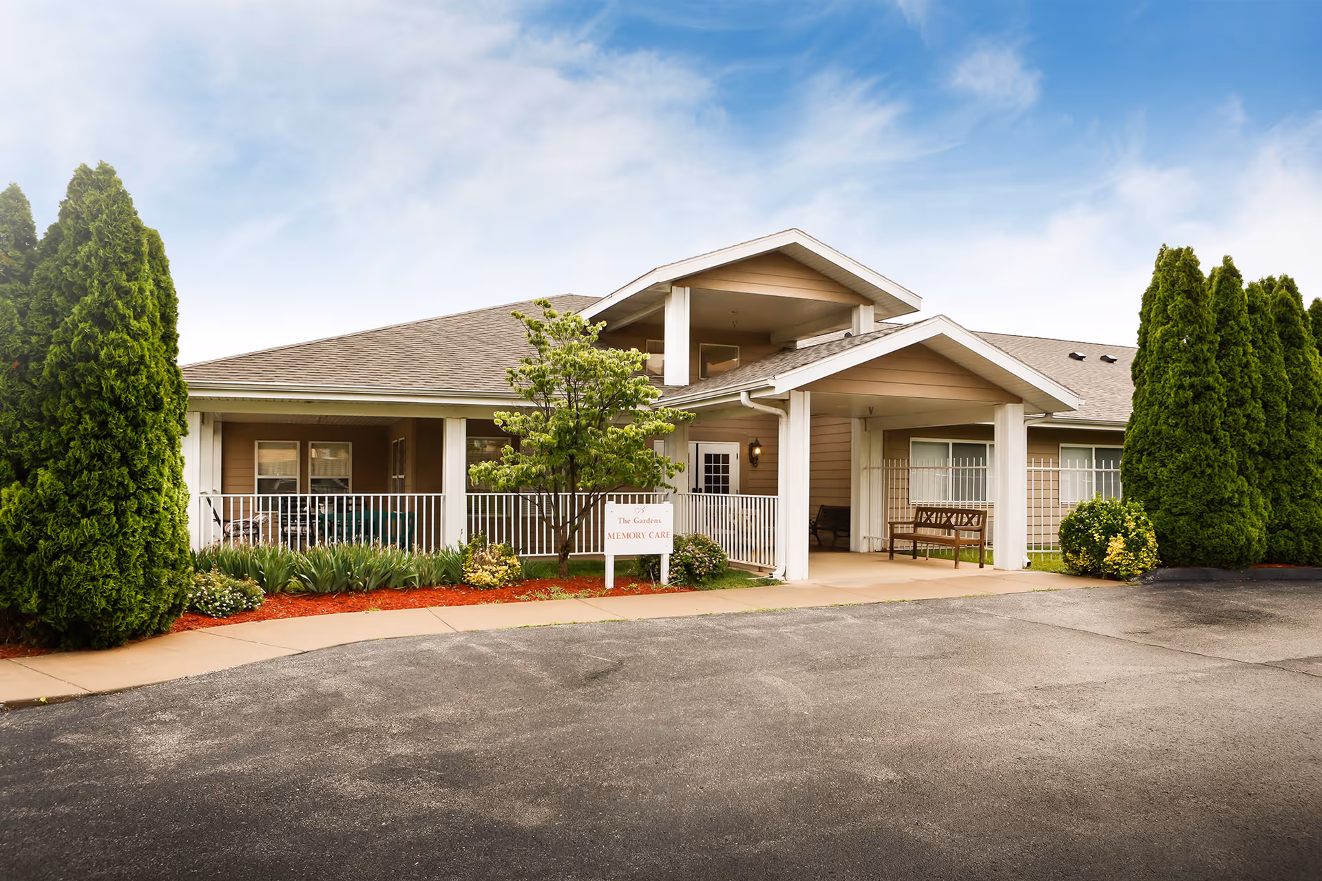 Exterior view of a single-story building with a covered entrance, surrounded by greenery including tall bushes and a small tree. There is a sign near the entrance that reads 'The Gardens Memory Care'. The sky is partly cloudy.