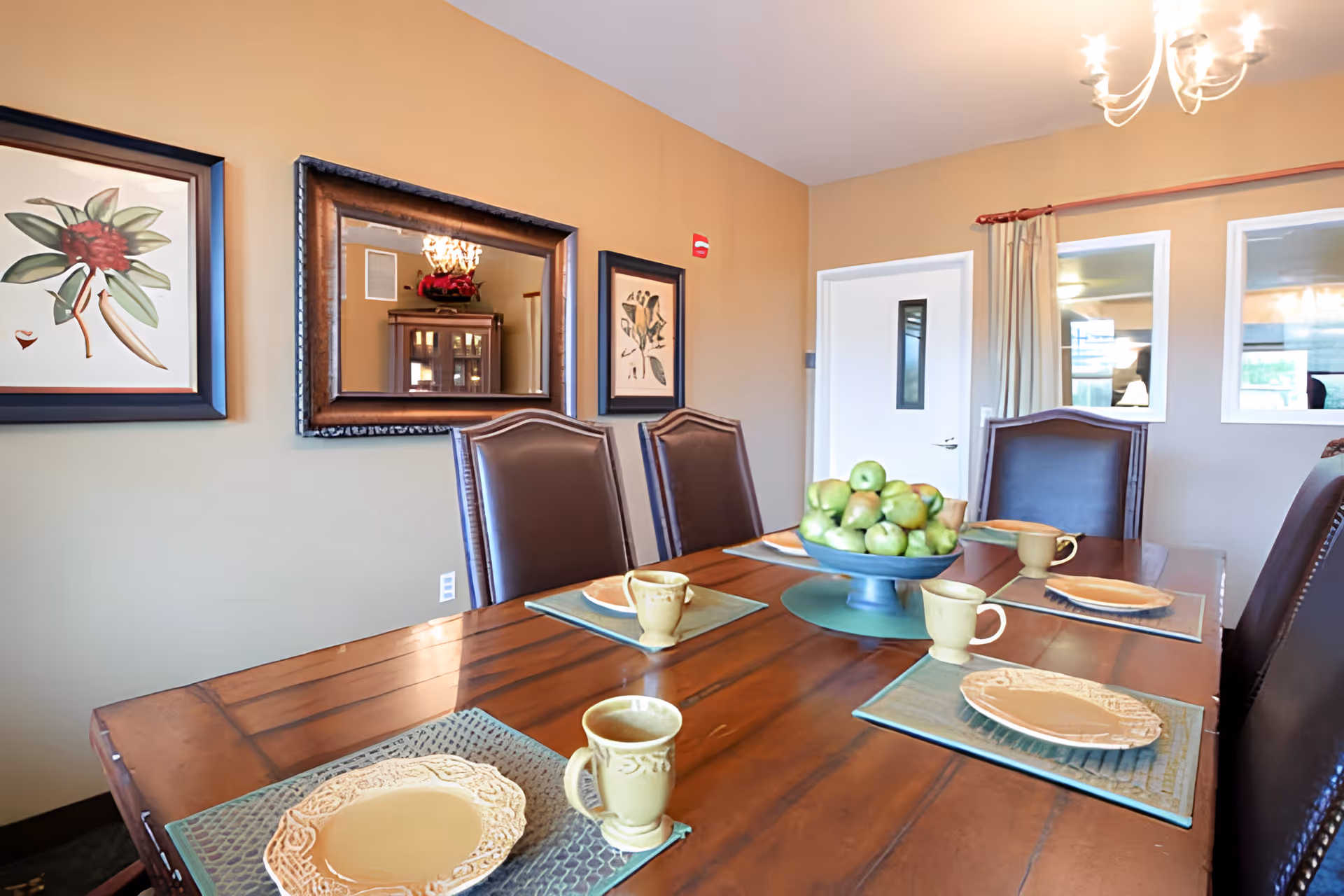 A dining room with a wooden table set with plates, mugs, placemats and a bowl of green apples, surrounded by leather chairs and framed wall art.