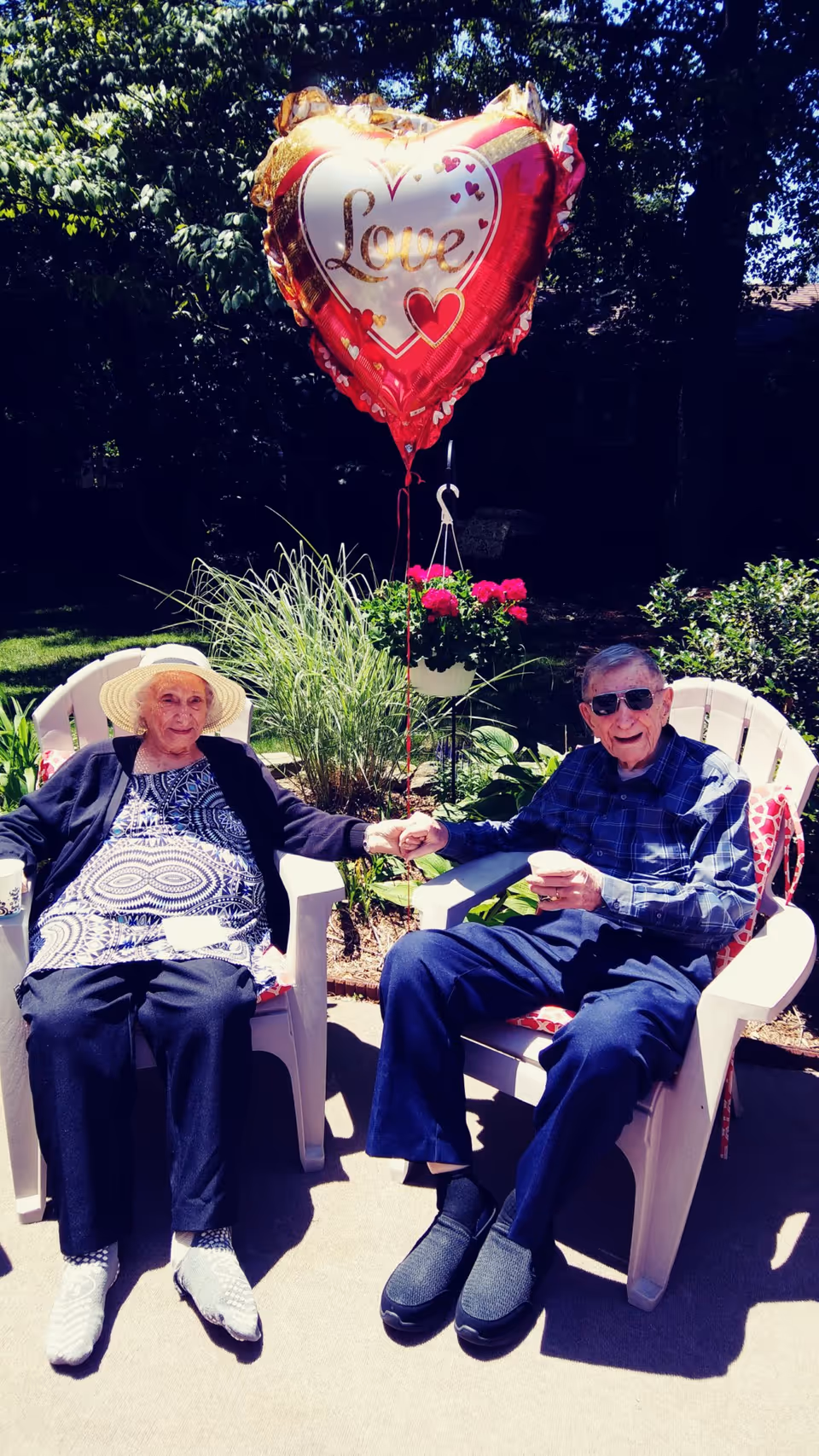 An elderly couple sitting outdoors in white plastic chairs holding hands. The woman is wearing a wide-brimmed hat, a patterned blouse, and a black cardigan. The man is wearing sunglasses, a blue plaid shirt, and dark pants. Behind them is a garden with green plants and a hanging pot of pink flowers. Above them is a heart-shaped balloon with the word 'Love' written on it.