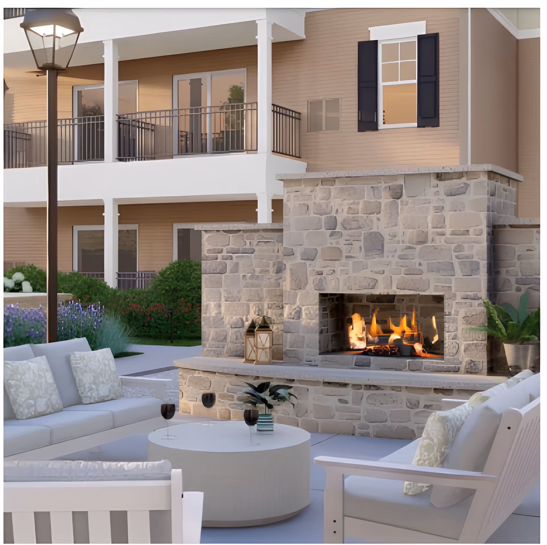 Outdoor seating area with cushioned white chairs and a round white table in front of a stone fireplace with a fire burning. The background shows a two-story building with balconies, windows, and greenery including bushes and flowers.
