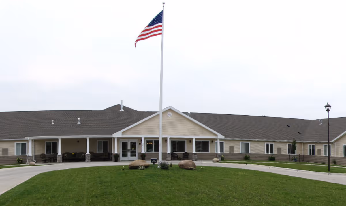 Single-story senior living facility with a covered main entrance, a tall flagpole flying an American flag, and a circular driveway surrounded by lawn.