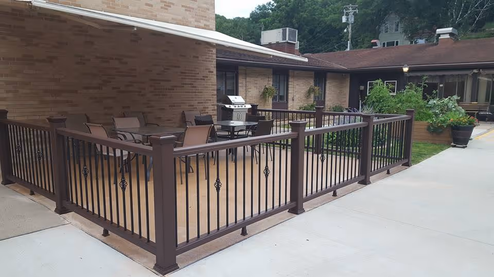 Outdoor patio area at St Michael's Assisted Living with a brown metal railing enclosing a concrete space furnished with a table, several chairs, and a barbecue grill. The patio is adjacent to a brick building with windows and hanging plants, and there are garden planters with greenery nearby.