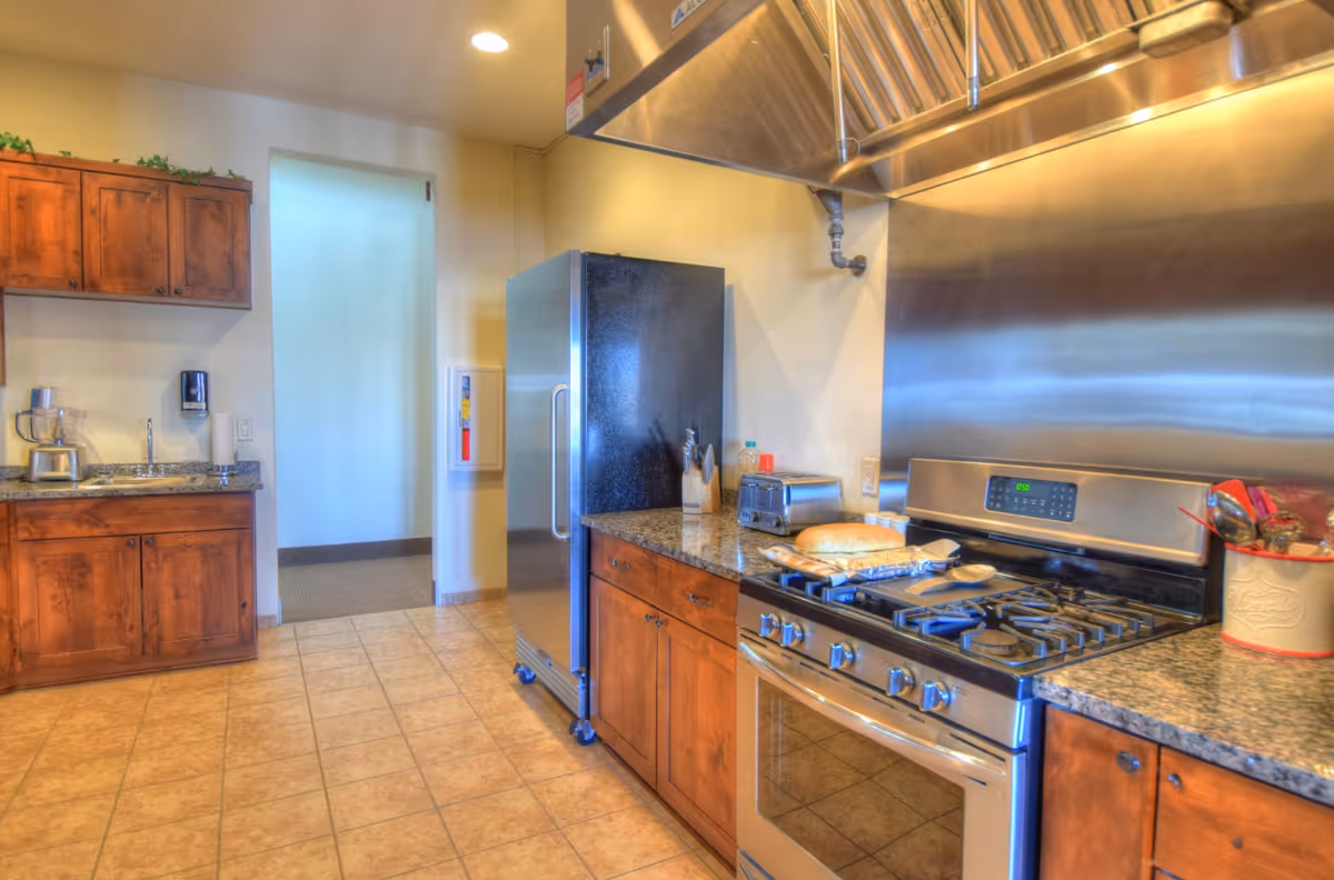 Commercial-style kitchen with a stainless steel stove and hood, refrigerator, wooden cabinets, and tiled floor.