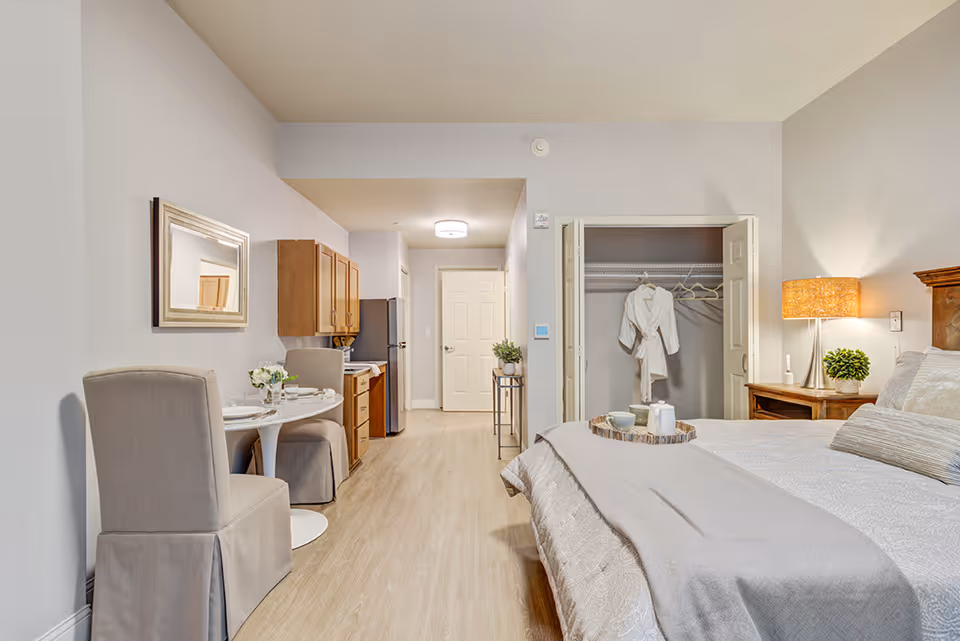 A cozy senior living studio apartment featuring a bed with a gray blanket and pillows, a wooden nightstand with a lamp and a small plant, an open closet with a white robe hanging inside, a small dining table with two upholstered chairs, a kitchenette with wooden cabinets, a refrigerator, and a mirror on the wall. The room has light-colored walls and wood flooring.
