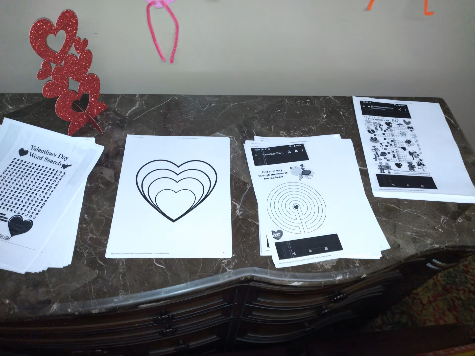 A dark marble countertop with several printed Valentine's Day activity sheets including a word search, a heart coloring page, a maze, and a crossword puzzle. There is a red glittery decoration with multiple hearts on the left side of the countertop.
