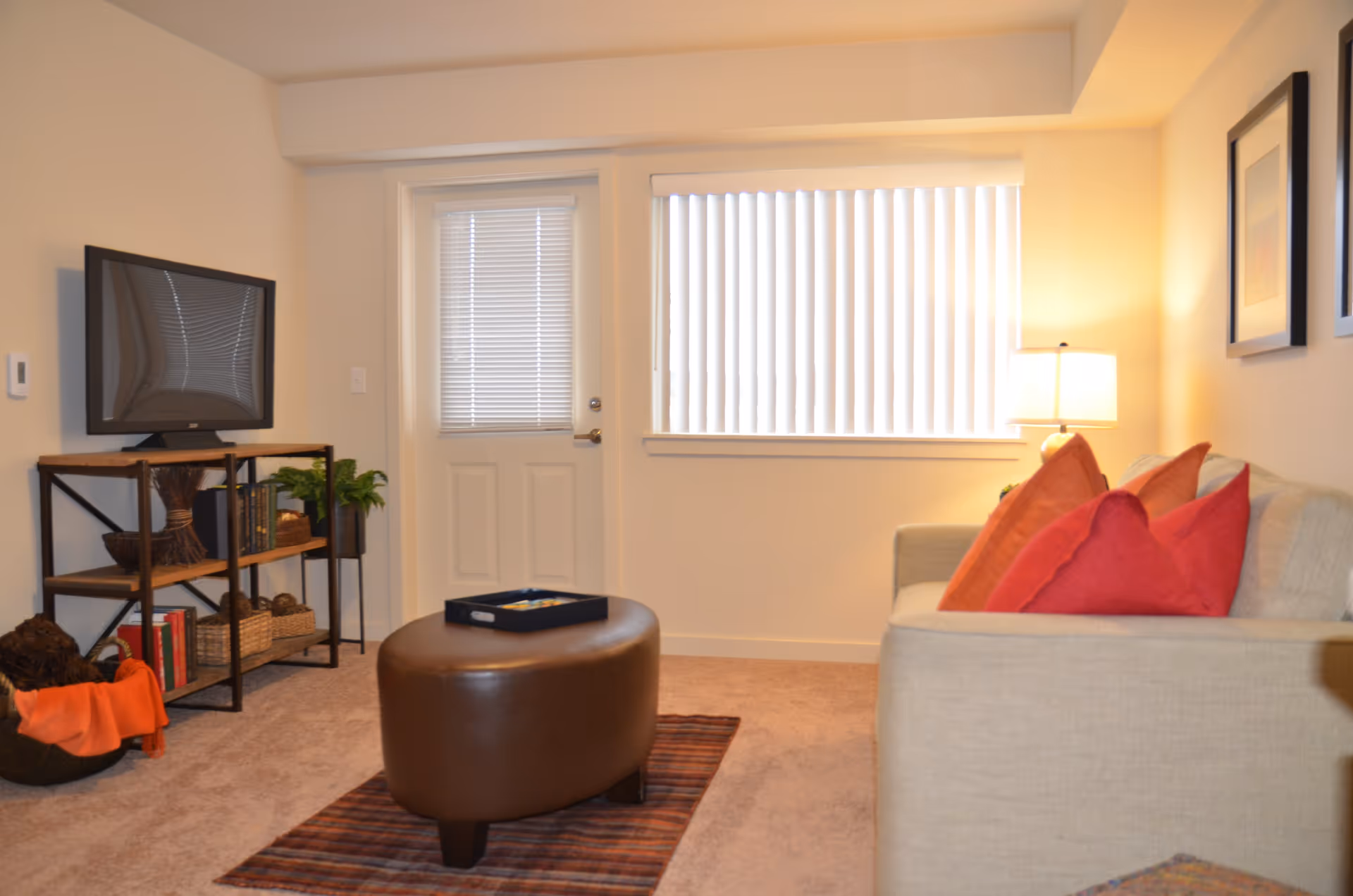 Cozy living room with a beige sofa with red pillows, a brown ottoman on a rug, a TV on shelving, and a door and window with blinds.