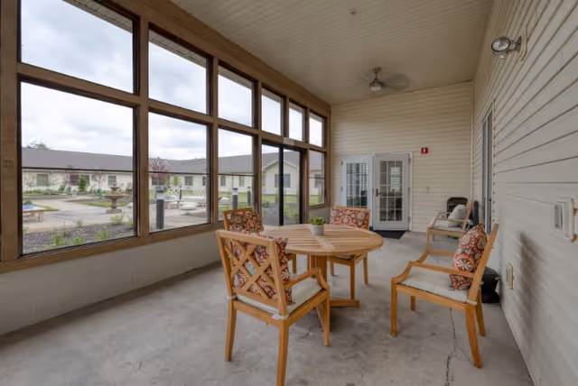 A covered outdoor patio area with a round wooden table surrounded by four wooden chairs with patterned cushions. The patio has large screened windows providing a view of a courtyard with a fountain and buildings in the background. The ceiling is white with a ceiling fan, and there is a door leading inside the building.