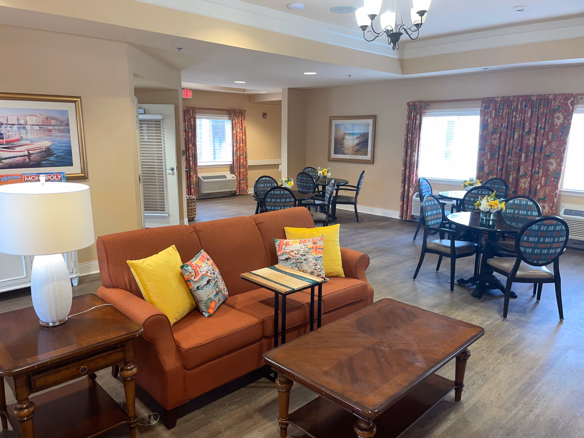 Community lounge featuring an orange sofa with colorful pillows and a coffee table in the foreground and round dining tables with chairs in the background.