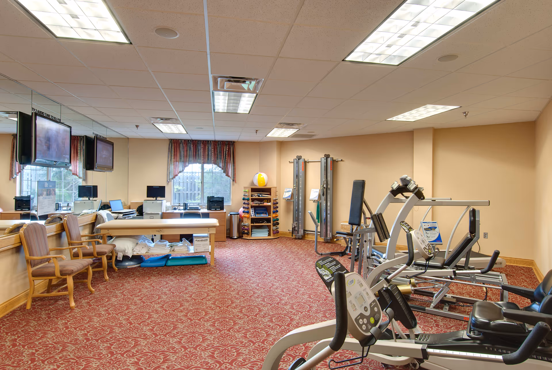 A spacious fitness and therapy room with exercise equipment including stationary bikes and weight machines. The room has a patterned red carpet, beige walls, and a ceiling with fluorescent lights. There are two wooden chairs, a treatment table with pillows and folded towels underneath, and a shelf with exercise balls and other fitness accessories. Two large windows with red and white curtains allow natural light in, and two wall-mounted TVs are visible on the left side.