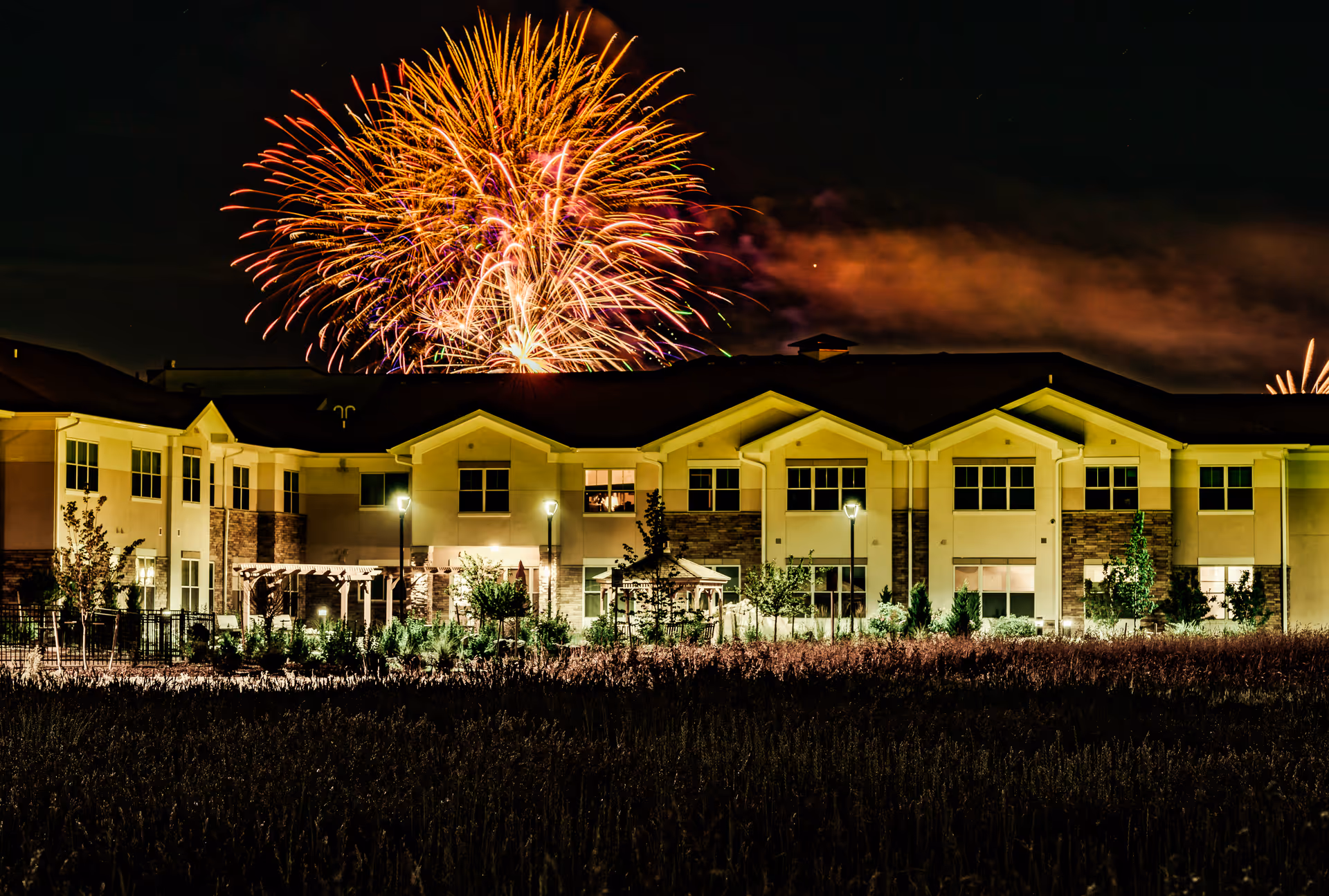 Nighttime view of a two-story assisted living facility building with exterior lights on and fireworks exploding in the sky behind it.