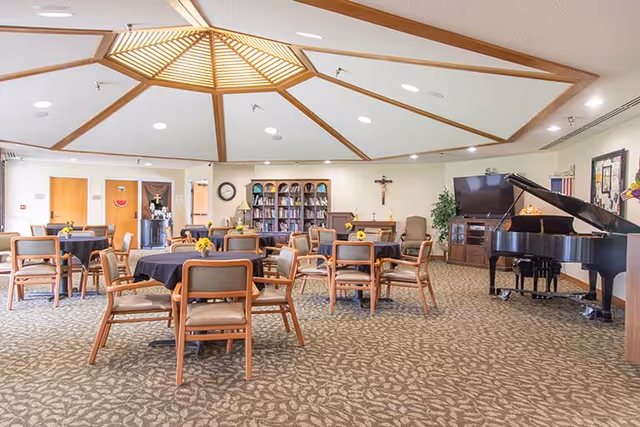 Spacious communal dining/activity room with round tables and chairs, a grand piano, bookshelf and decorative ceiling feature.