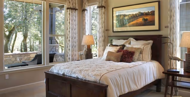 A cozy bedroom with a large bed featuring white and beige bedding and multiple pillows. There are two bedside tables with lamps on each side of the bed. A framed landscape painting hangs above the bed. Large windows with patterned curtains allow natural light to fill the room and offer a view of an outdoor patio area with trees and stone walls.