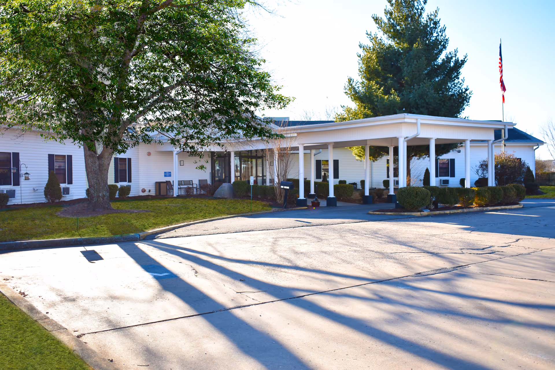 Exterior view of Prairie Grove Health & Rehab building with a covered entrance, surrounded by trees and shrubs, and an American flag on a flagpole to the right.