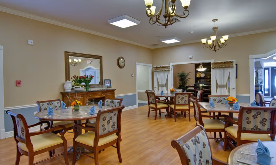 A bright communal dining room with round tables set with napkins and small floral centerpieces and upholstered chairs.