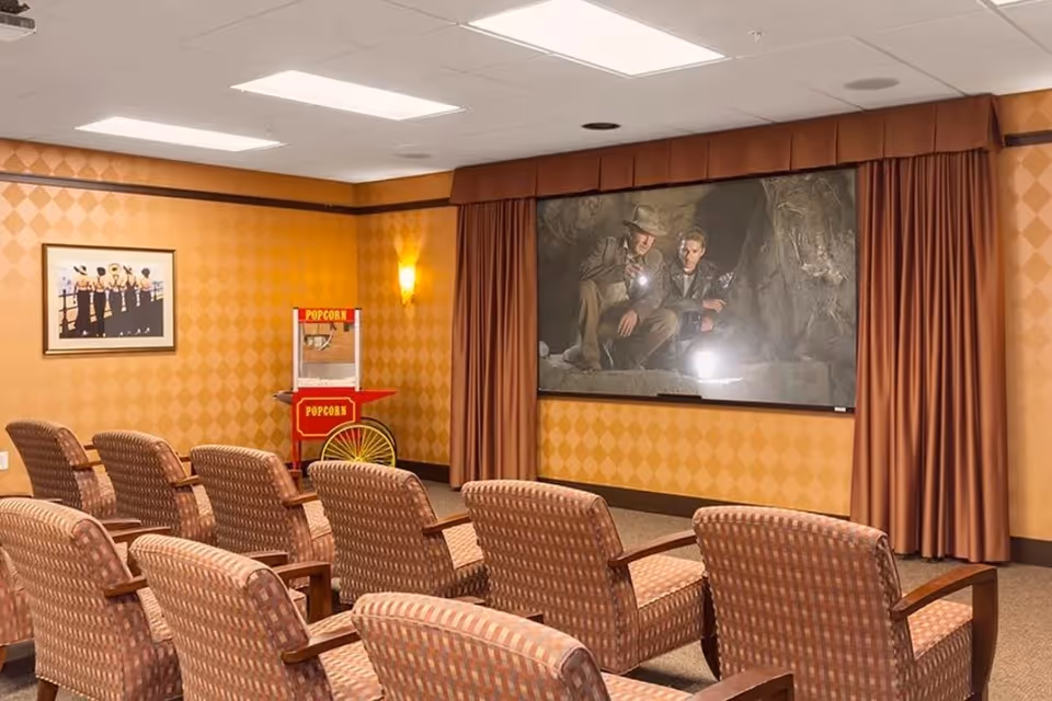 Small screening room with rows of upholstered chairs facing a large screen, curtains, and a popcorn cart.