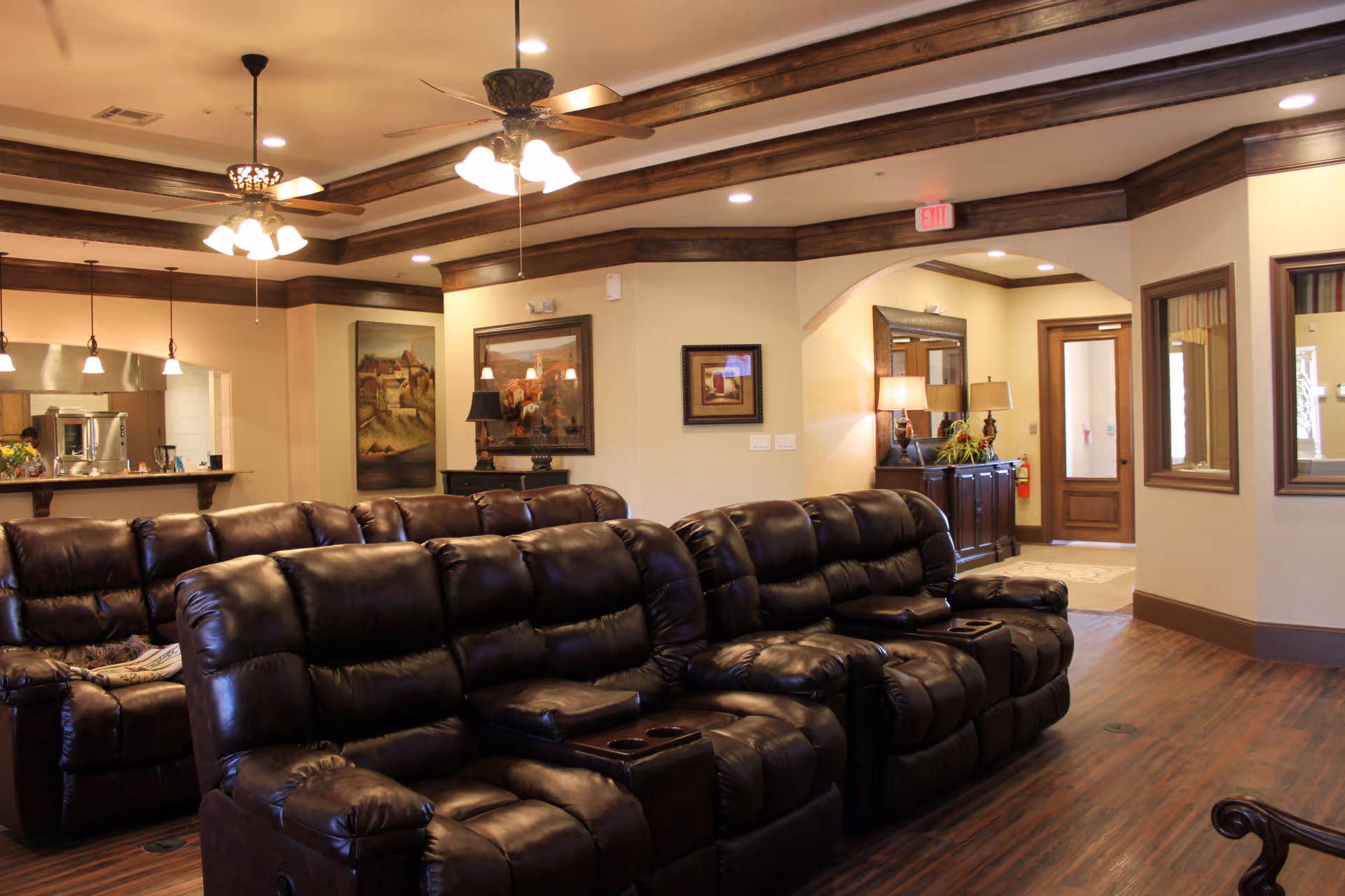 Interior view of a comfortable living room area with multiple dark brown leather recliner sofas arranged in rows. The room features wooden flooring, ceiling fans with lights, framed artwork on the walls, and a sideboard with lamps and plants. The background shows an open kitchen area with hanging pendant lights and a doorway leading to another room.