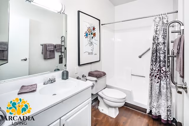 A clean and modern bathroom featuring a white vanity with a sink, a large mirror above it, a toilet with folded towels on top, and a bathtub with a floral patterned shower curtain. The walls are white, and there is a framed artwork of a hand holding colorful flowers above the toilet. Towels hang on a rack and a door handle is visible on the right side.