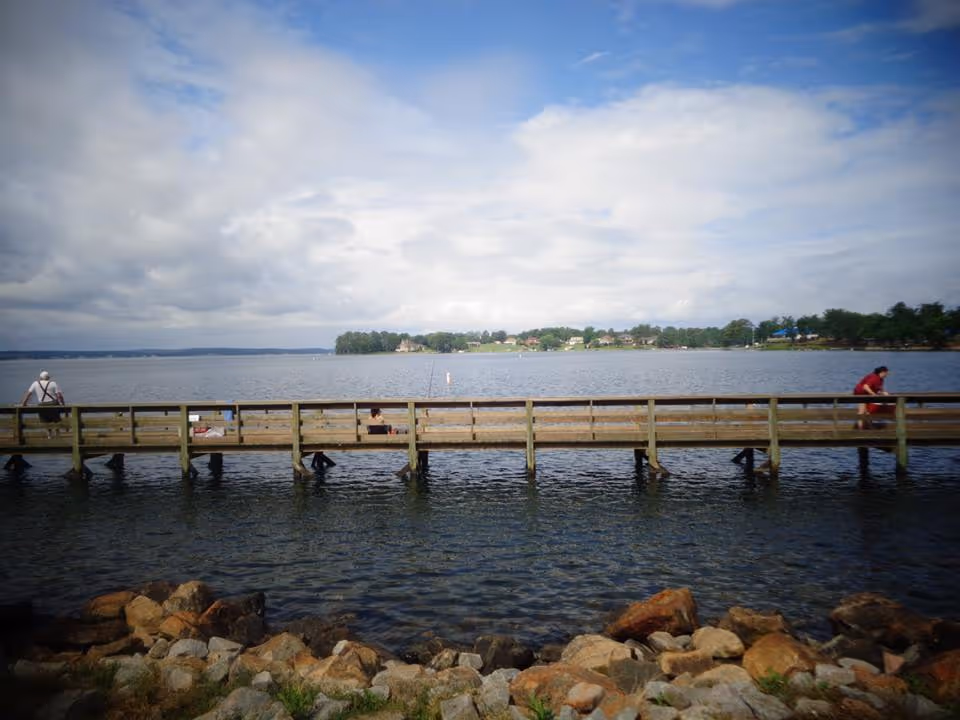 A wooden pier extending over a body of water with two people sitting at opposite ends. The sky is partly cloudy and there are trees and houses visible in the distance across the water. Rocks line the shore in the foreground.
