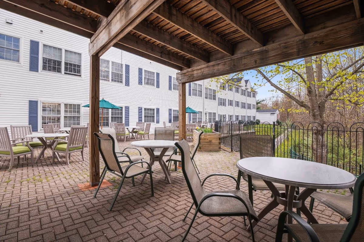 Outdoor patio area at Middlebrook Farms at Trumbull with multiple round tables and chairs arranged on a brick-paved surface. The patio is partially covered by a wooden pergola and overlooks a fenced garden area with trees and greenery. The white building with multiple windows and blue shutters is visible in the background.